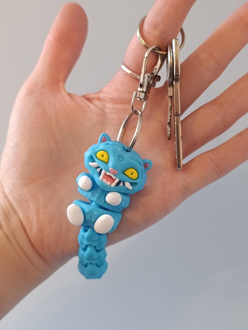 May include: A blue cat-shaped keyring with yellow eyes and white teeth. The keyring is attached to a silver clasp and ring, with several keys hanging from the ring. The cat has a segmented body and white paws.