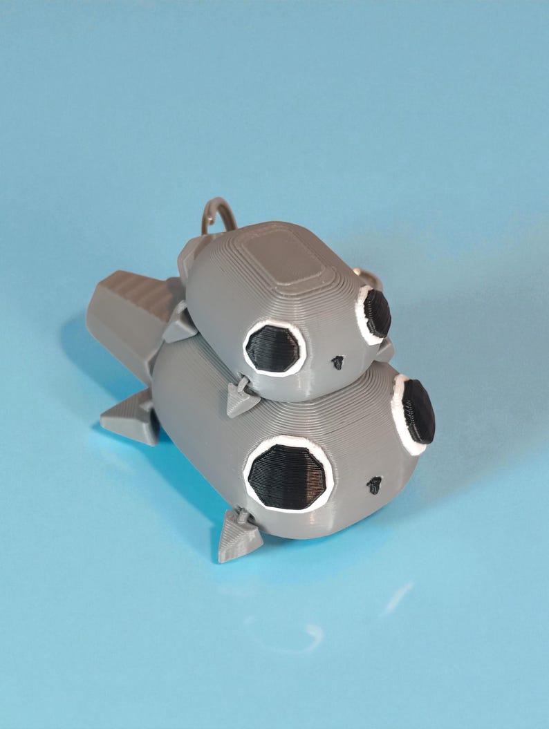 May include: A gray 3D-printed keychain featuring a stacked design of two cartoon-like creatures. Each creature has large black eyes with white outlines and a small, smiling mouth. The keychain has a silver metal ring.