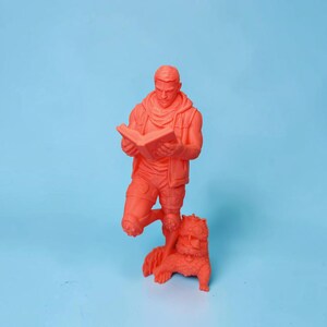 May include: A monochromatic neon orange figurine depicting a standing figure wearing a hooded vest and shorts while reading an open book. A small, furry creature with large eyes and textured fur sits at the feet of the figure, looking upward.