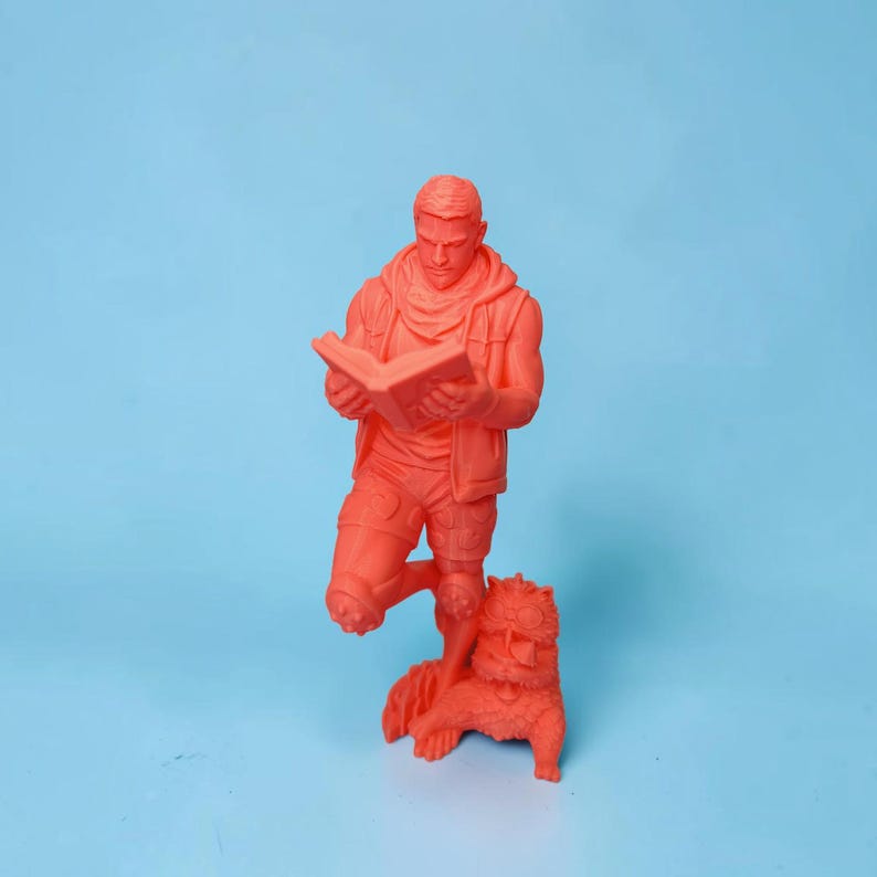 May include: A monochromatic neon orange figurine depicting a standing figure wearing a hooded vest and shorts while reading an open book. A small, furry creature with large eyes and textured fur sits at the feet of the figure, looking upward.
