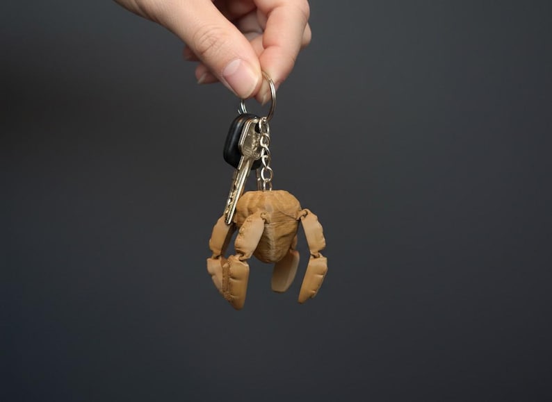 May include: A wooden articulated figure keychain with a key ring and keys. The figure is light brown and has a rounded head and jointed limbs. The background is a solid dark gray.