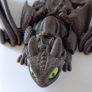K&ouml;nnte beinhalten: 3D-gedruckte schwarze Drachenfigur mit leuchtend gr&uuml;nen Augen. Der Drache hat detaillierte Fl&uuml;gel, einen strukturierten K&ouml;rper und einen detaillierten Kopf. Die Figur liegt, mit dem Kopf zum Betrachter.