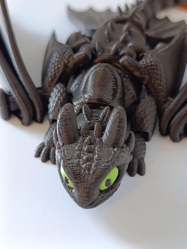 K&ouml;nnte beinhalten: 3D-gedruckte schwarze Drachenfigur mit leuchtend gr&uuml;nen Augen. Der Drache hat detaillierte Fl&uuml;gel, einen strukturierten K&ouml;rper und einen detaillierten Kopf. Die Figur liegt, mit dem Kopf zum Betrachter.