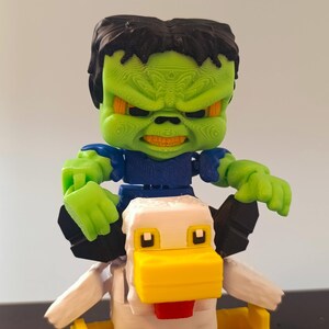 May include: A 3D-printed toy featuring a green-skinned figure with black hair and a stern expression, riding a white and yellow chicken. The figure is wearing a blue shirt and has green hands. The chicken has a yellow beak and red tongue.