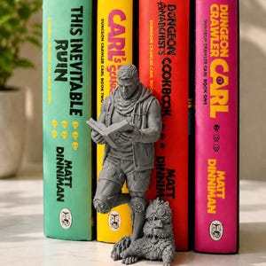May include: A gray resin bookend featuring a standing figure reading a book, wearing a hooded vest and heart-patterned shorts, with a small furry creature wearing goggles at their feet, positioned in front of four Dungeon Crawler Carl books.