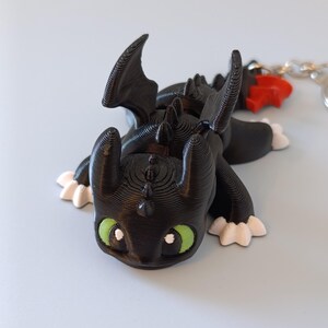 May include: A black dragon figurine with green eyes and white accents on its feet. The dragon has large wings and a detailed, textured surface. A red charm and a silver chain are attached to the figurine, suggesting it may be a keychain.