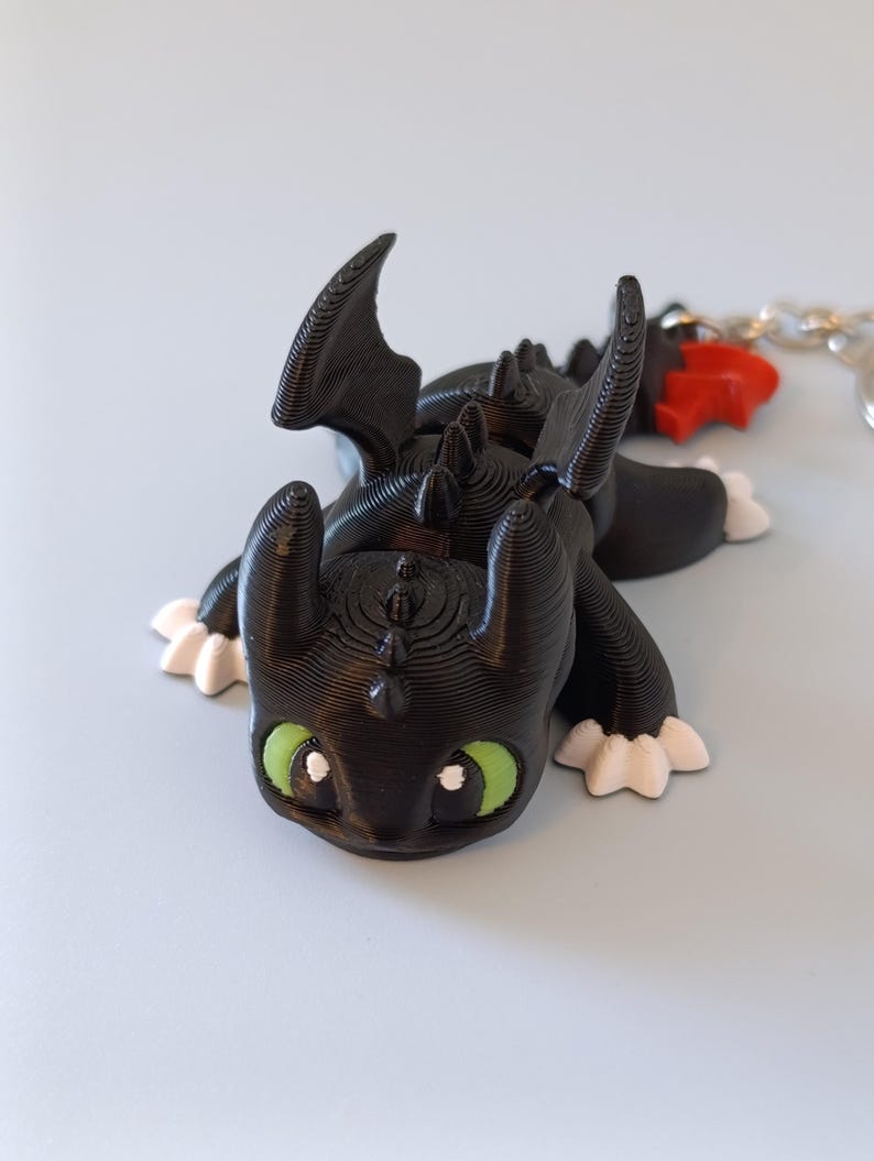 May include: A black dragon figurine with green eyes and white accents on its feet. The dragon has large wings and a detailed, textured surface. A red charm and a silver chain are attached to the figurine, suggesting it may be a keychain.