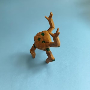 Pode incluir: Uma figura estilizada de cor castanha de uma criatura com corpo arredondado e m&uacute;ltiplos membros. A figura tem manchas verdes e est&aacute; posada com os bra&ccedil;os levantados. O fundo &eacute; azul claro liso.
