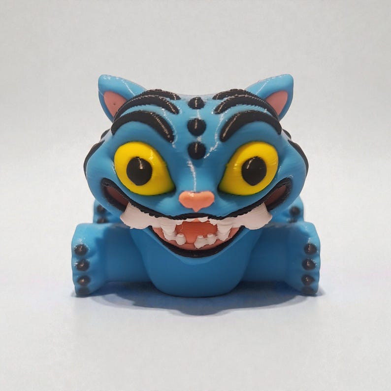 Puede incluir: Una figura de gato azul con grandes ojos amarillos, una nariz rosada y una amplia sonrisa con dientes. El gato tiene rayas y manchas negras, y sus orejas son rosas. La figura es un dise&ntilde;o caprichoso de dibujos animados.