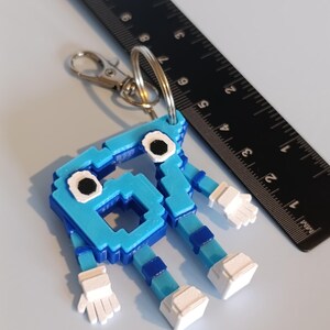 Pode incluir: Chaveiro azul pixelizado em forma de personagem de desenho animado com m&atilde;os e p&eacute;s brancos. Possui um mosquet&atilde;o e um porta-chaves prateados. Uma r&eacute;gua preta est&aacute; no fundo. Altura aproximada: 7 cm.