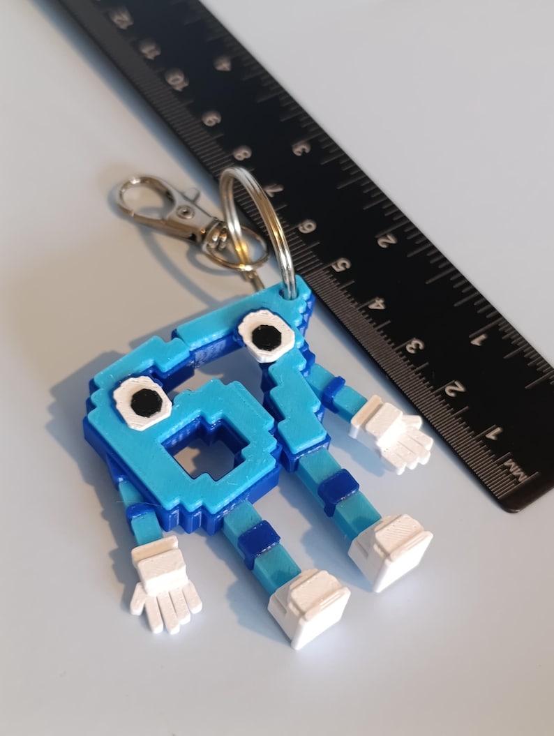 Pode incluir: Chaveiro azul pixelizado em forma de personagem de desenho animado com m&atilde;os e p&eacute;s brancos. Possui um mosquet&atilde;o e um porta-chaves prateados. Uma r&eacute;gua preta est&aacute; no fundo. Altura aproximada: 7 cm.
