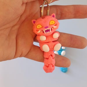 May include: A pink cat-shaped keyring with yellow eyes and white teeth. The keyring is attached to a silver metal clasp. The cat's body is segmented, and it has white paws. A blue and white keyring is visible in the background.