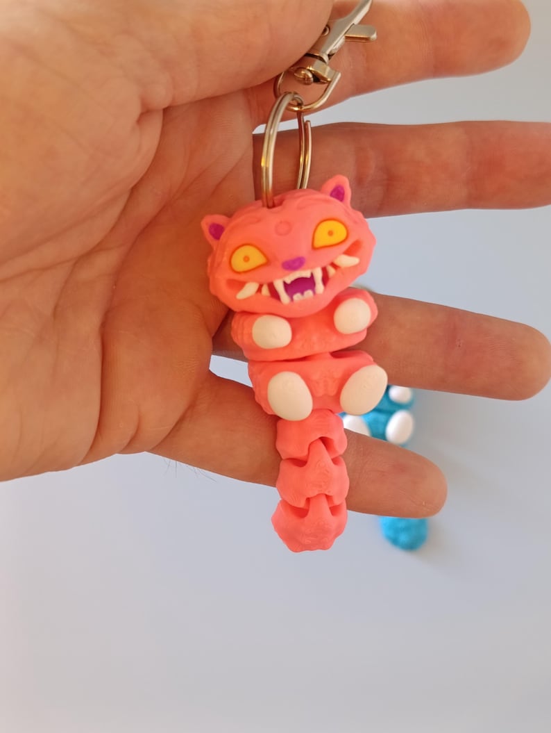 May include: A pink cat-shaped keyring with yellow eyes and white teeth. The keyring is attached to a silver metal clasp. The cat's body is segmented, and it has white paws. A blue and white keyring is visible in the background.
