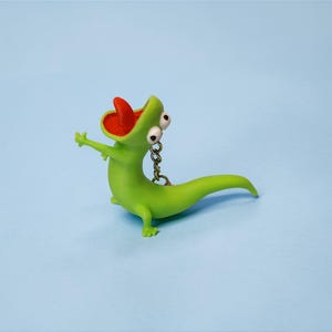 May include: A green lizard keychain with a red mouth and a surprised expression. The lizard has large, googly eyes and outstretched arms. The keychain is made of a smooth material and has a silver chain.