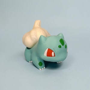 May include: A teal Bulbasaur figurine with red eyes and green spots, carrying a white bulb on its back. The figurine is a whimsical representation of the popular Pok&eacute;mon character, perfect for collectors or fans of the series.