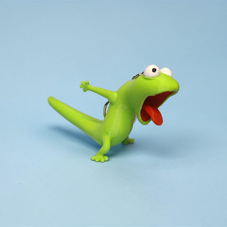 May include: A green lizard-shaped keychain with a wide open mouth and a red tongue. The lizard has white eyes and is posed with its arms outstretched. The keychain is on a blue background.