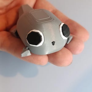 May include: A gray 3D-printed figurine of a stylized creature with large black eyes, white outlines, and small fins. The figurine is held in a hand, showcasing its details and texture.