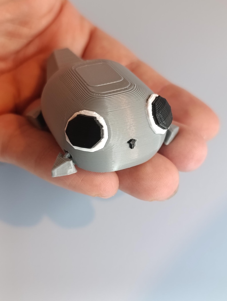 May include: A gray 3D-printed figurine of a stylized creature with large black eyes, white outlines, and small fins. The figurine is held in a hand, showcasing its details and texture.