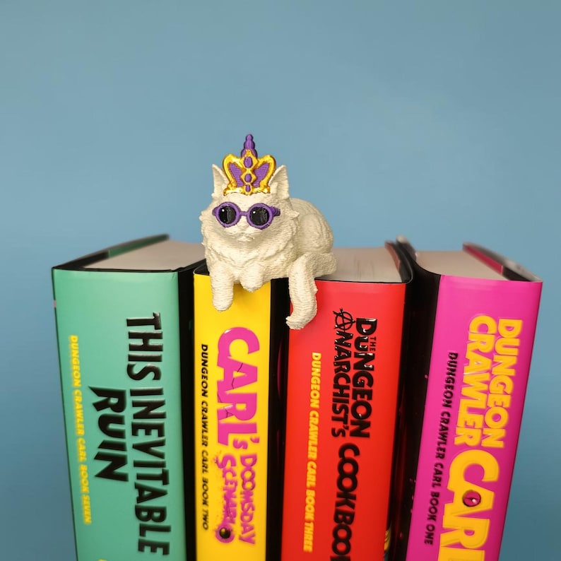 Op de afbeelding: Een wit kattenbeeldje met een paarse en gouden kroon en een paarse zonnebril, zittend op vier boeken met de titels This Inevitable Ruin, Carl's Doomsday Scenario, The Dungeon Crawler Narchist's Cookbook en Dungeon Crawler Carl.