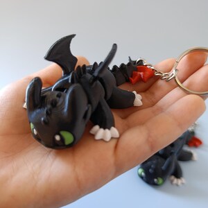 May include: Black dragon keychain with green eyes, white claws, and red accents. The dragon has a detailed, textured design and is attached to a silver keyring. The dragon is lying down, with its wings spread.