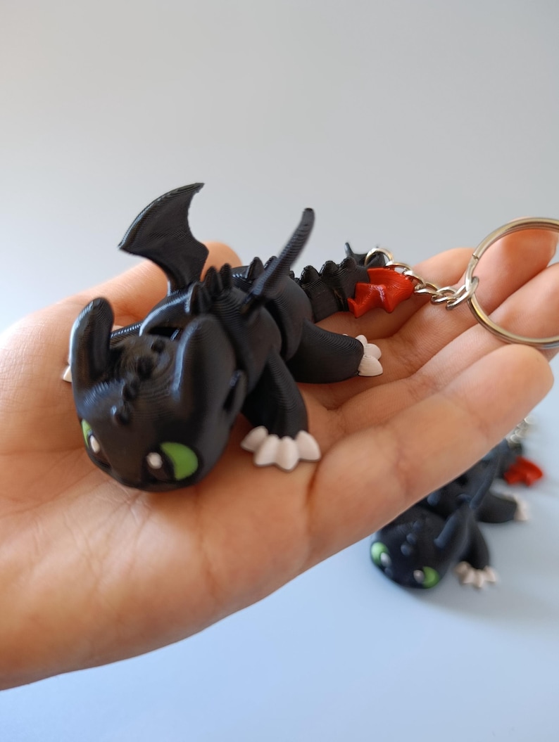 May include: Black dragon keychain with green eyes, white claws, and red accents. The dragon has a detailed, textured design and is attached to a silver keyring. The dragon is lying down, with its wings spread.
