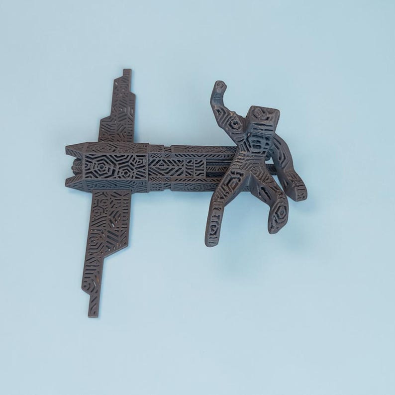 K&ouml;nnte beinhalten: Eine braune, 3D-gedruckte Skulptur mit einer stilisierten Figur und einer kreuzf&ouml;rmigen Struktur. Die Figur ist mit erhobenem Arm dargestellt, und beide Elemente weisen komplizierte geometrische Muster auf. Die Skulptur ist vor einem hellblauen Hintergrund platziert.