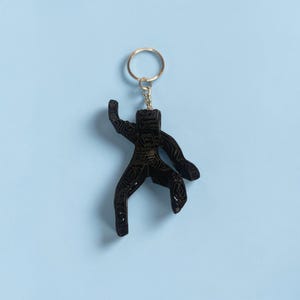 Puede incluir: Llavero negro con forma de figura estilizada con un anillo dorado. La figura tiene una superficie texturizada y est&aacute; suspendida sobre un fondo azul claro. El llavero es un accesorio decorativo.