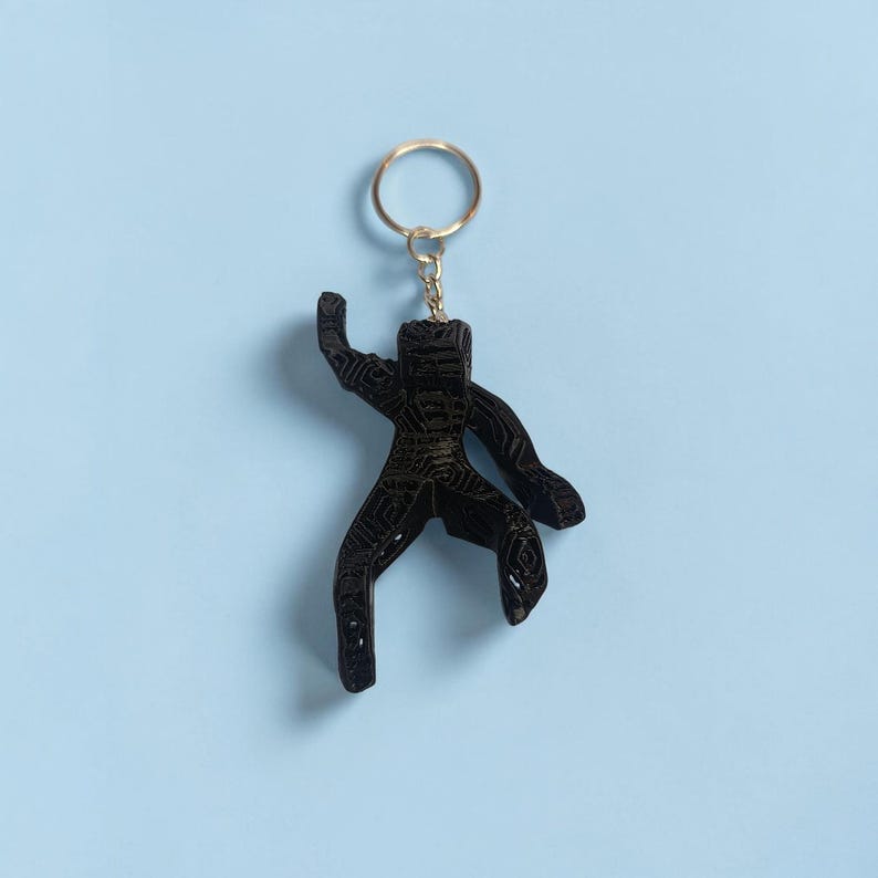 Puede incluir: Llavero negro con forma de figura estilizada con un anillo dorado. La figura tiene una superficie texturizada y est&aacute; suspendida sobre un fondo azul claro. El llavero es un accesorio decorativo.