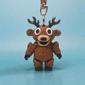 May include: A brown reindeer keychain with a silver clasp. The reindeer has large white eyes, a black nose, and small black antlers. The body is textured and the keychain is hanging against a light blue background.