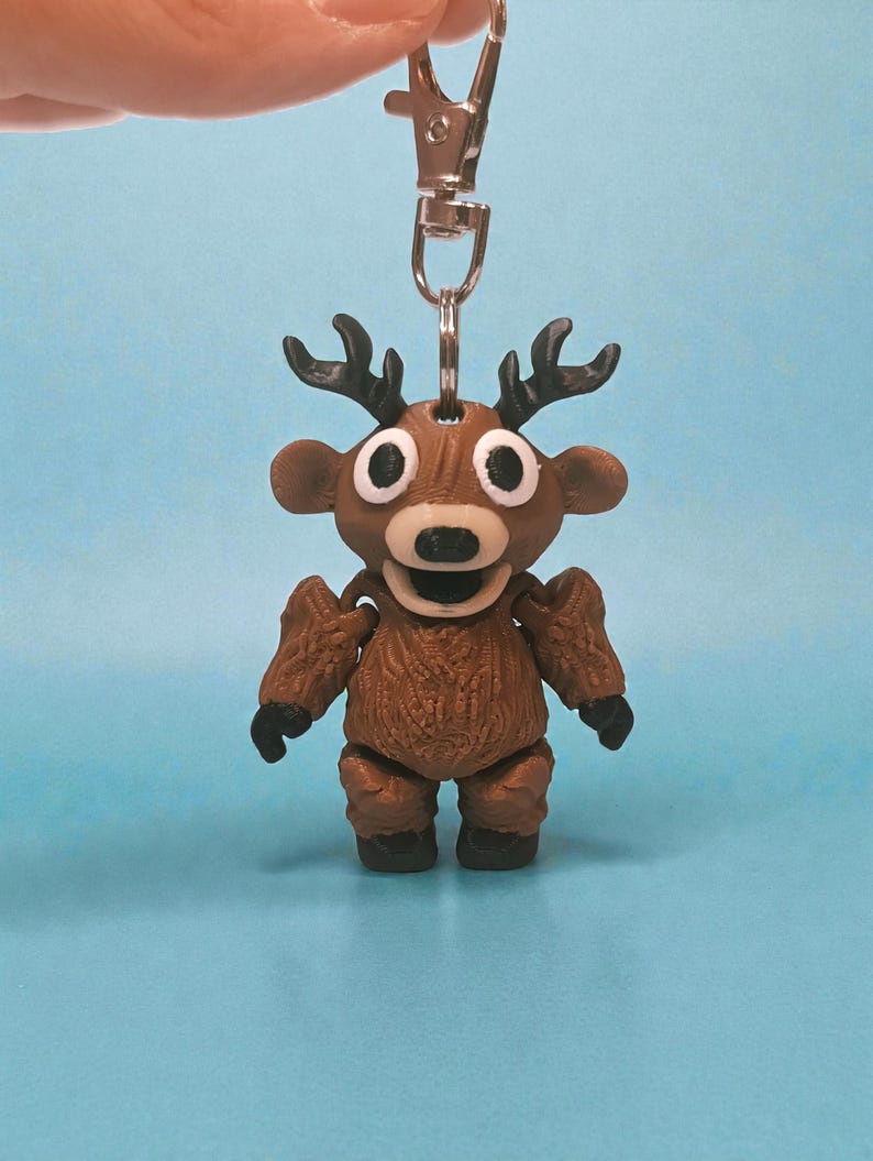 May include: A brown reindeer keychain with a silver clasp. The reindeer has large white eyes, a black nose, and small black antlers. The body is textured and the keychain is hanging against a light blue background.