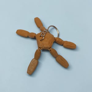 May include: A brown articulated wooden figure keychain with a silver keyring. The figure has movable arms and legs, and is posed in a star shape. The background is a solid light blue.