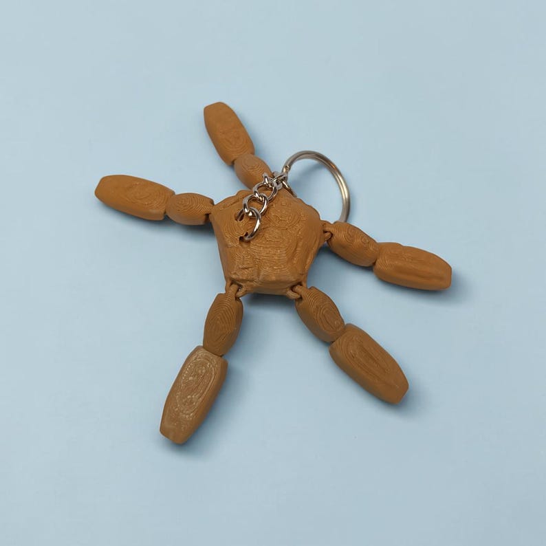 May include: A brown articulated wooden figure keychain with a silver keyring. The figure has movable arms and legs, and is posed in a star shape. The background is a solid light blue.
