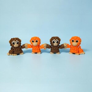 May include: Four small, colorful monkey figurines. Two are brown, and two are orange, all with large, expressive eyes. They are holding hands, creating a playful and friendly scene. The background is a solid light blue.