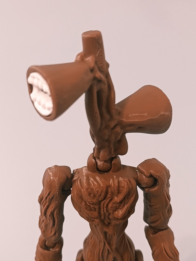 Op de afbeelding: Een bruin Siren Head actiefiguur met een gedetailleerd, gestructureerd ontwerp. De figuur heeft twee luidsprekerachtige hoofden met witte tanden, beweegbare armen en een menselijk torso. De figuur staat tegen een effen achtergrond.