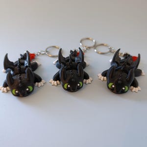 May include: Three black dragon keychains with green eyes, white paws, and red accents. The keychains are lying on a light blue surface. Each dragon has a silver keyring attached.