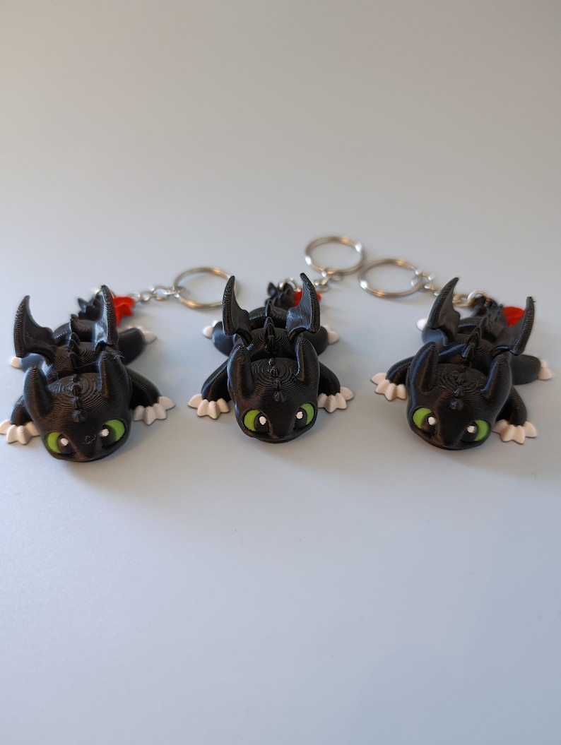 May include: Three black dragon keychains with green eyes, white paws, and red accents. The keychains are lying on a light blue surface. Each dragon has a silver keyring attached.