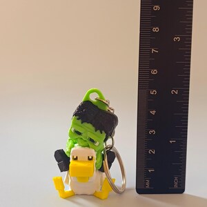 Op de afbeelding: Een novelty sleutelhanger met een groene Frankenstein-figuur die op een gele en witte eend rijdt. De sleutelhanger heeft een metalen ring en is ongeveer 5 cm hoog.