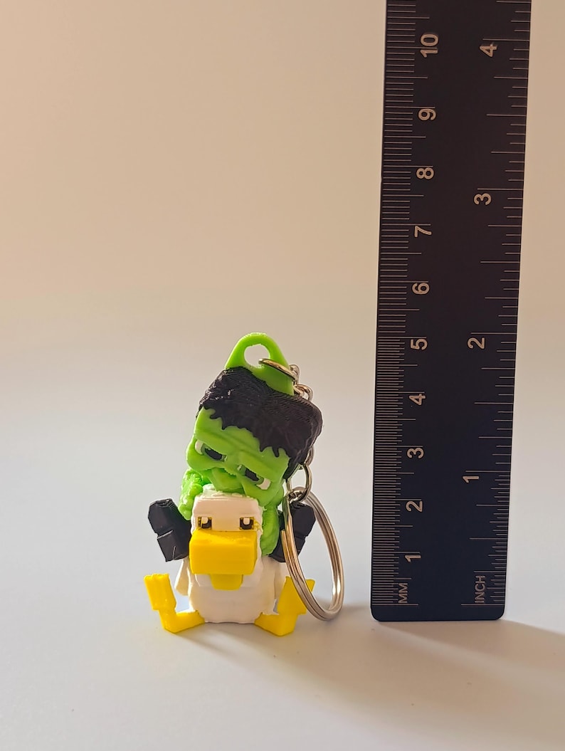 Op de afbeelding: Een novelty sleutelhanger met een groene Frankenstein-figuur die op een gele en witte eend rijdt. De sleutelhanger heeft een metalen ring en is ongeveer 5 cm hoog.