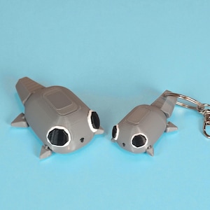 May include: Two gray, 3D-printed fish-shaped keychains with black and white details. One keychain is attached to a silver keyring. The fish have a rectangular body, fins, and a tail. The background is a solid light blue.