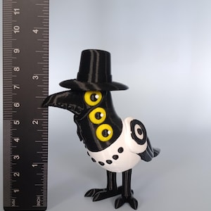 Puede incluir: Una caprichosa figura de cuervo en blanco y negro con sombrero de copa y tres ojos amarillos. El cuervo mide aproximadamente 10 cm de altura, con una regla en el fondo para la escala. El dise&ntilde;o del ave es &uacute;nico y de dibujos animados.