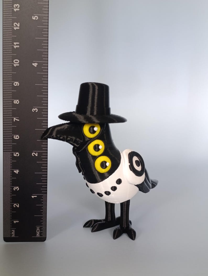Puede incluir: Una caprichosa figura de cuervo en blanco y negro con sombrero de copa y tres ojos amarillos. El cuervo mide aproximadamente 10 cm de altura, con una regla en el fondo para la escala. El dise&ntilde;o del ave es &uacute;nico y de dibujos animados.