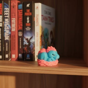 Puede incluir: Una peque&ntilde;a figura impresa en 3D de una criatura durmiente con cuerpo azul claro y crin rosa, que descansa dentro de una base rosa texturizada en forma de nido. El objeto est&aacute; colocado sobre un estante de madera frente a varios libros.