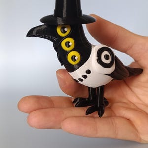 Puede incluir: Una figura impresa en 3D de un p&aacute;jaro con sombrero de copa negro, pico y patas. El p&aacute;jaro tiene tres ojos amarillos y un cuerpo de estilo esmoquin blanco y negro. La figura se sostiene en una mano.