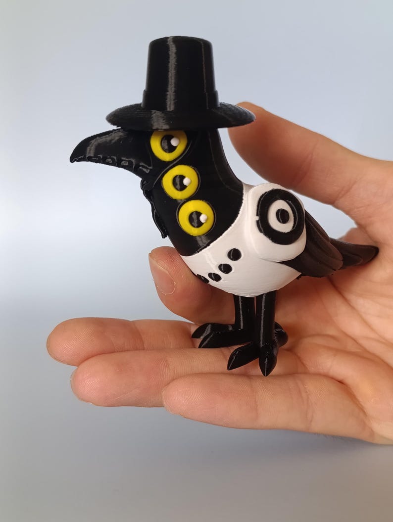 Puede incluir: Una figura impresa en 3D de un p&aacute;jaro con sombrero de copa negro, pico y patas. El p&aacute;jaro tiene tres ojos amarillos y un cuerpo de estilo esmoquin blanco y negro. La figura se sostiene en una mano.