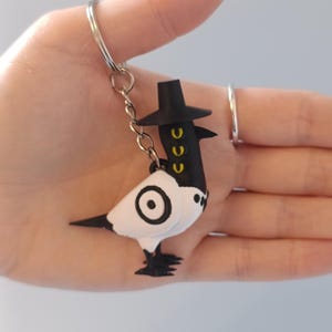 May include: A whimsical keychain featuring a black and white bird-like figure with a top hat. The bird has a circular design on its body and yellow accents. The keychain is attached to a silver ring.