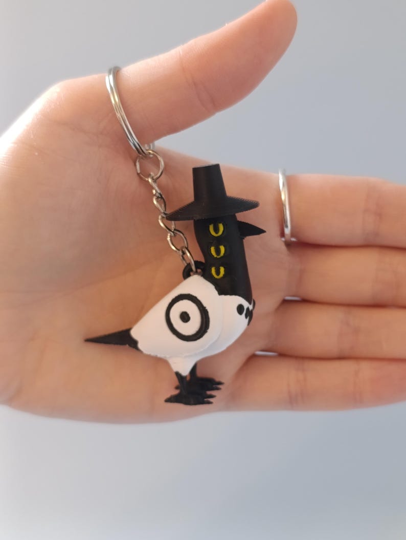 May include: A whimsical keychain featuring a black and white bird-like figure with a top hat. The bird has a circular design on its body and yellow accents. The keychain is attached to a silver ring.