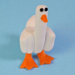 May include: A white goose figurine with orange feet and beak, and black eyes. The goose is standing on a light blue surface. The figurine has a cartoonish design.