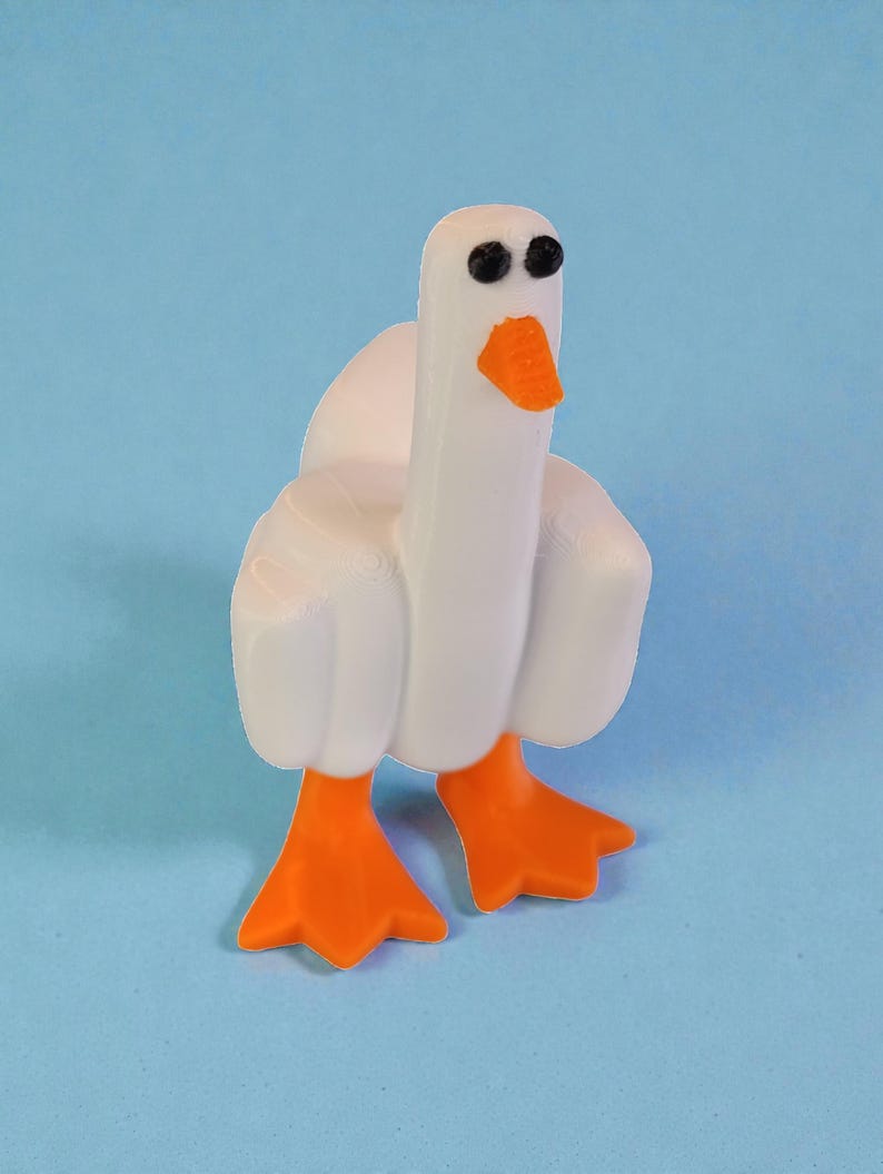 May include: A white goose figurine with orange feet and beak, and black eyes. The goose is standing on a light blue surface. The figurine has a cartoonish design.