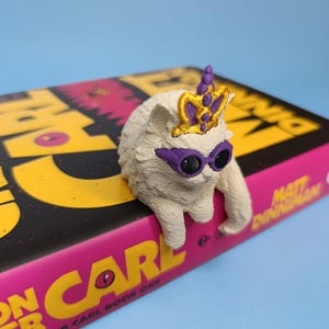 Op de afbeelding: Een klein, gebroken wit, getextureerd kattenbeeldje met een paarse zonnebril en een goudkleurige en paarse eenhoornhoorn. De kat zit op de rand van een boek met de titel Dungeon Crawler Carl van Matt Dinniman met gele tekst.