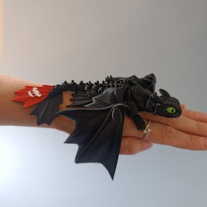 K&ouml;nnte beinhalten: Schwarze Drachenfigur mit gr&uuml;nen Augen und roter und wei&szlig;er Schwanzflosse. Der Drache hat detaillierte Fl&uuml;gel und ist auf einer Hand platziert. Das Objekt hat ein Fantasy-Thema.
